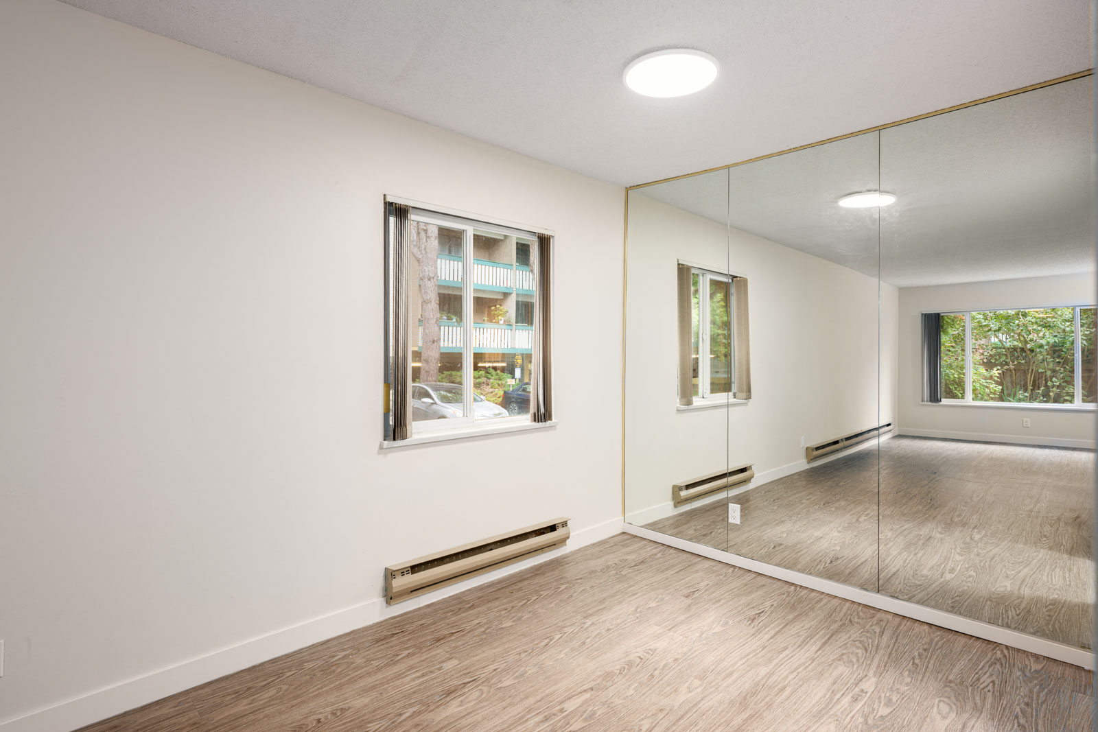 Empty room with light-colored walls, wood flooring, a large mirror wall, a window, and a baseboard heater below the window.