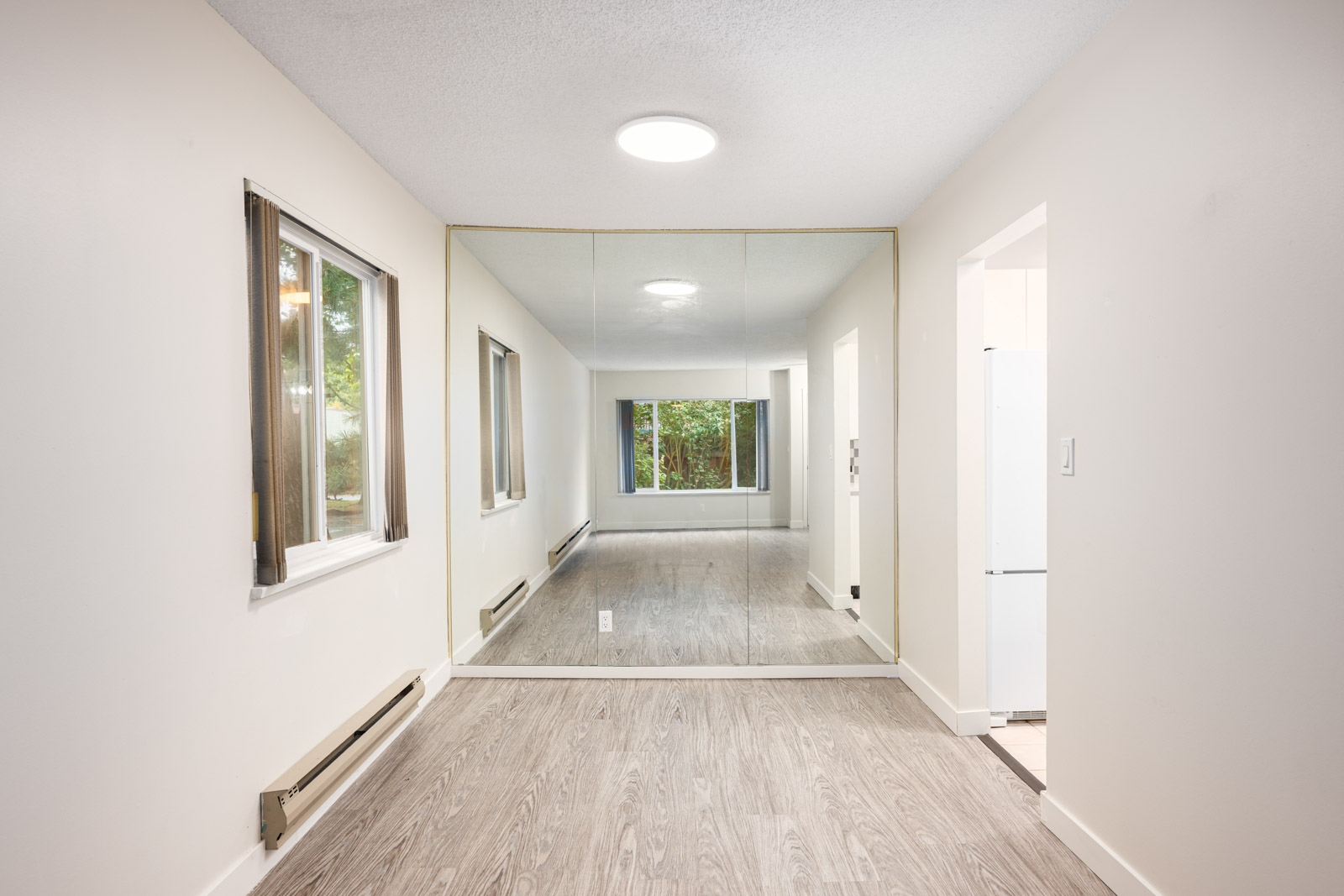 A narrow room with light-colored walls, wood-style flooring, a large wall mirror, two windows, and a glimpse of a kitchen area with a refrigerator.
