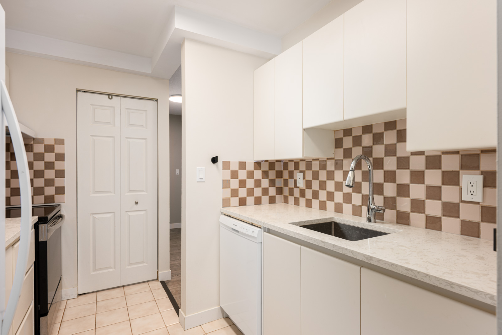 Modern kitchen with white cabinets, beige checkered tile backsplash, stainless steel sink, dishwasher, and closed double doors in the background.