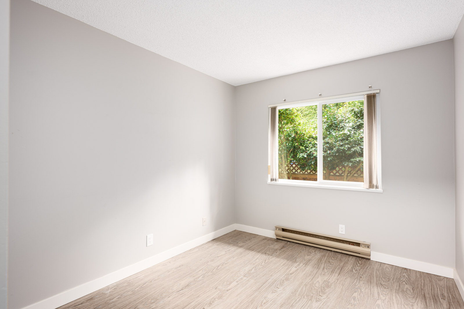 Empty room with light gray walls, wood-style flooring, a window with partial blinds, and a baseboard heater beneath the window.