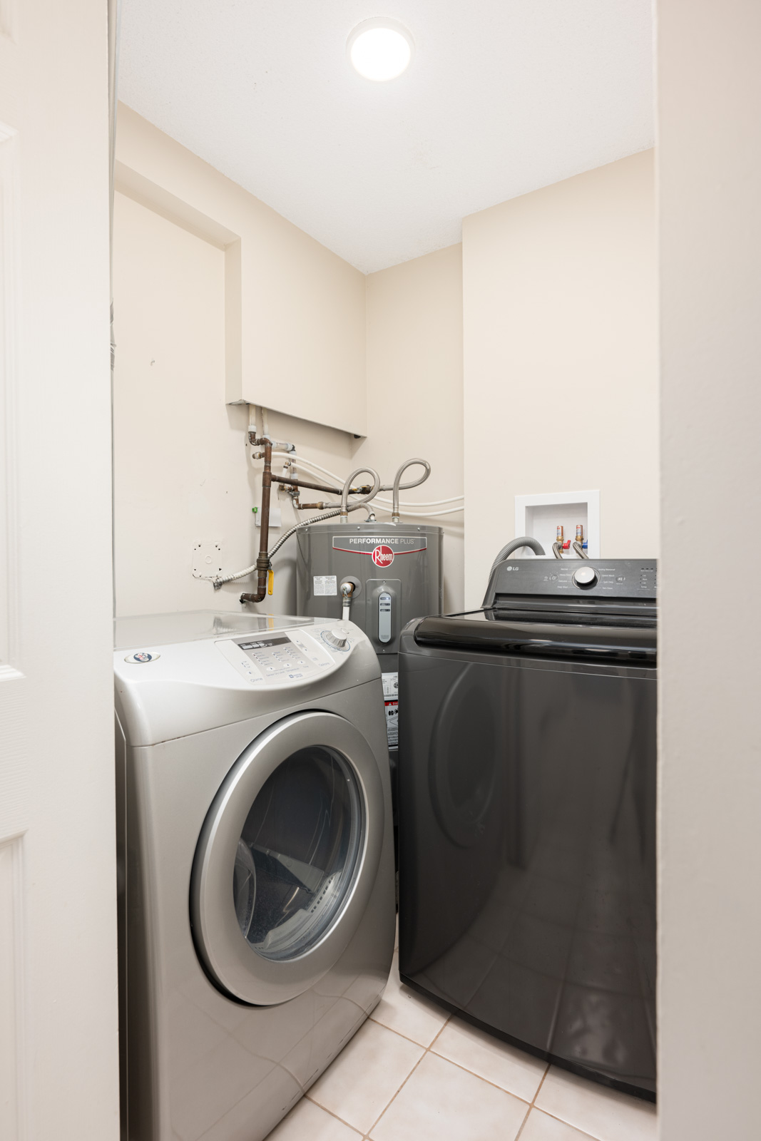 A small laundry room with a front-loading washing machine, a top-loading dryer, a water heater, and tiled flooring under bright ceiling light.