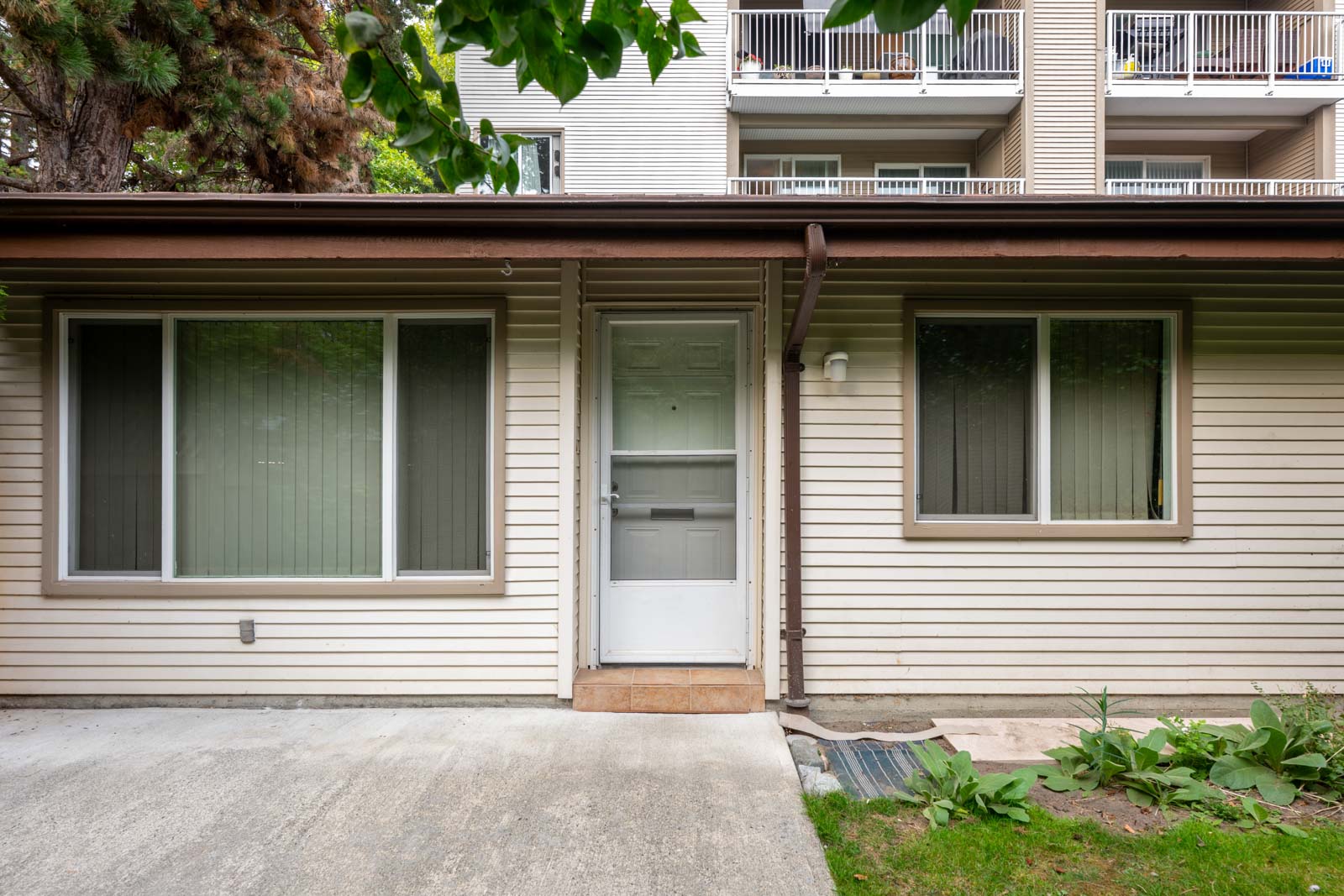 A beige single-story apartment entrance with a white door and two windows, surrounded by greenery and a concrete walkway, with balconies visible above.