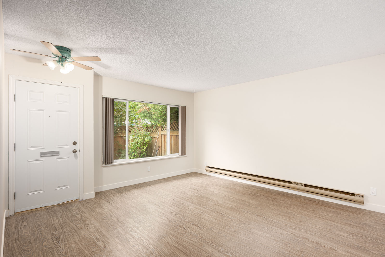 Empty room with light-colored walls, wood-style flooring, a large window, heater along the baseboard, and a white entrance door with a ceiling fan overhead.
