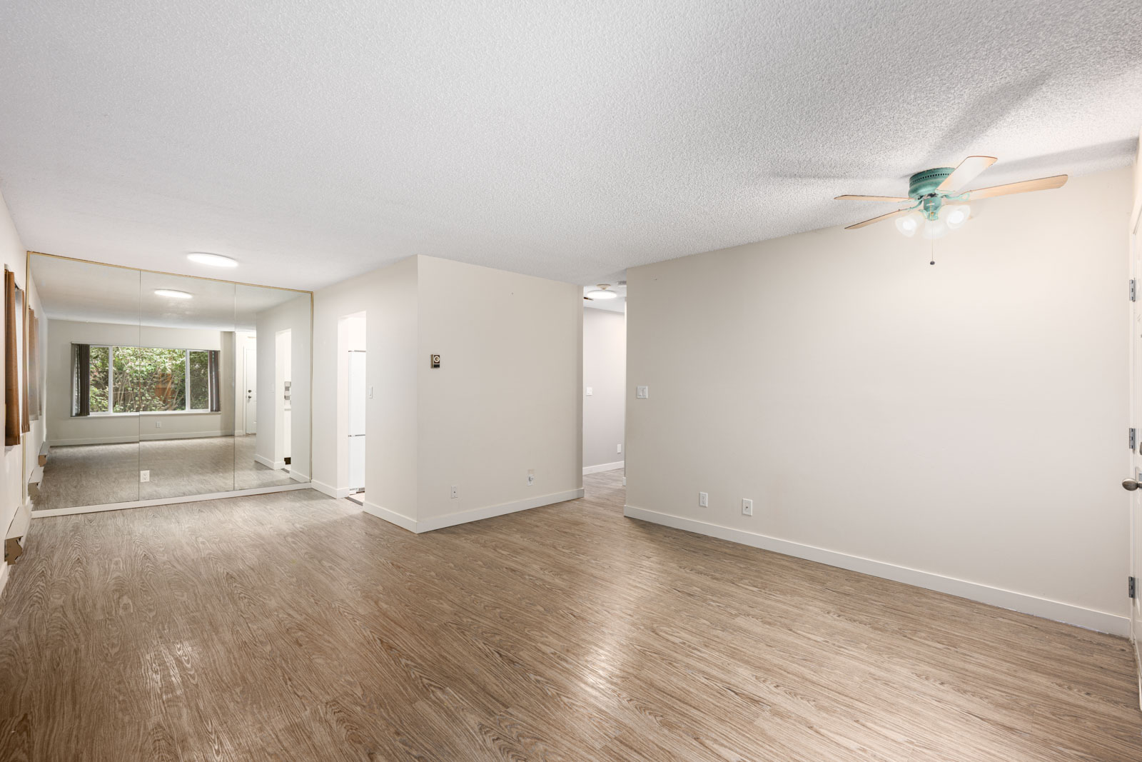 Unfurnished living room with wood flooring, white walls, a ceiling fan, mirrored closet doors, and a window overlooking greenery.