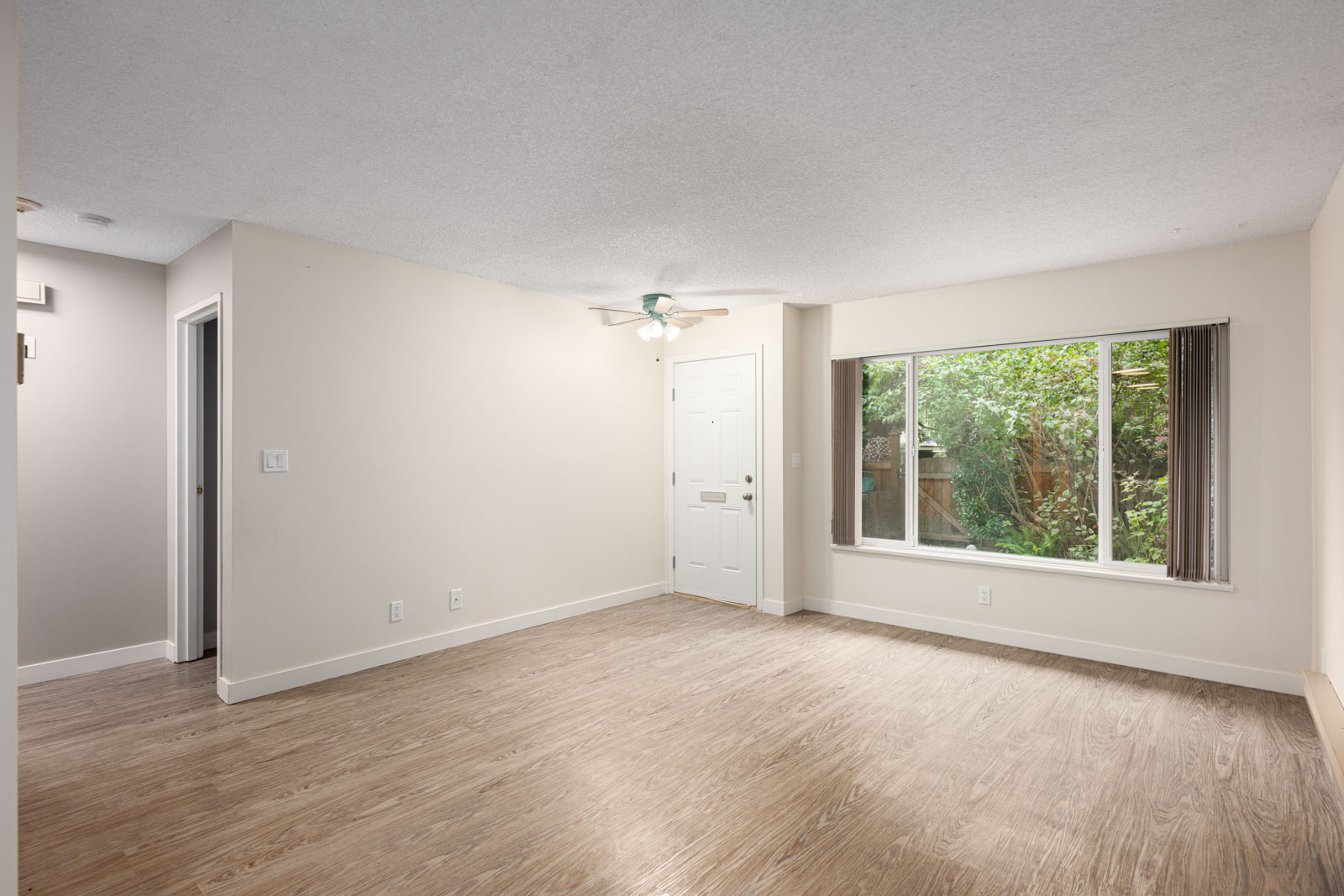 Unfurnished room with light wood flooring, beige walls, a large window with vertical blinds, a ceiling fan with lights, and a white front door.