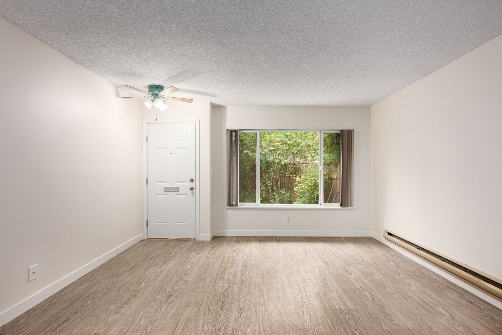Empty room with light wood flooring, white walls, a ceiling fan, large window with garden view, and a white front door.