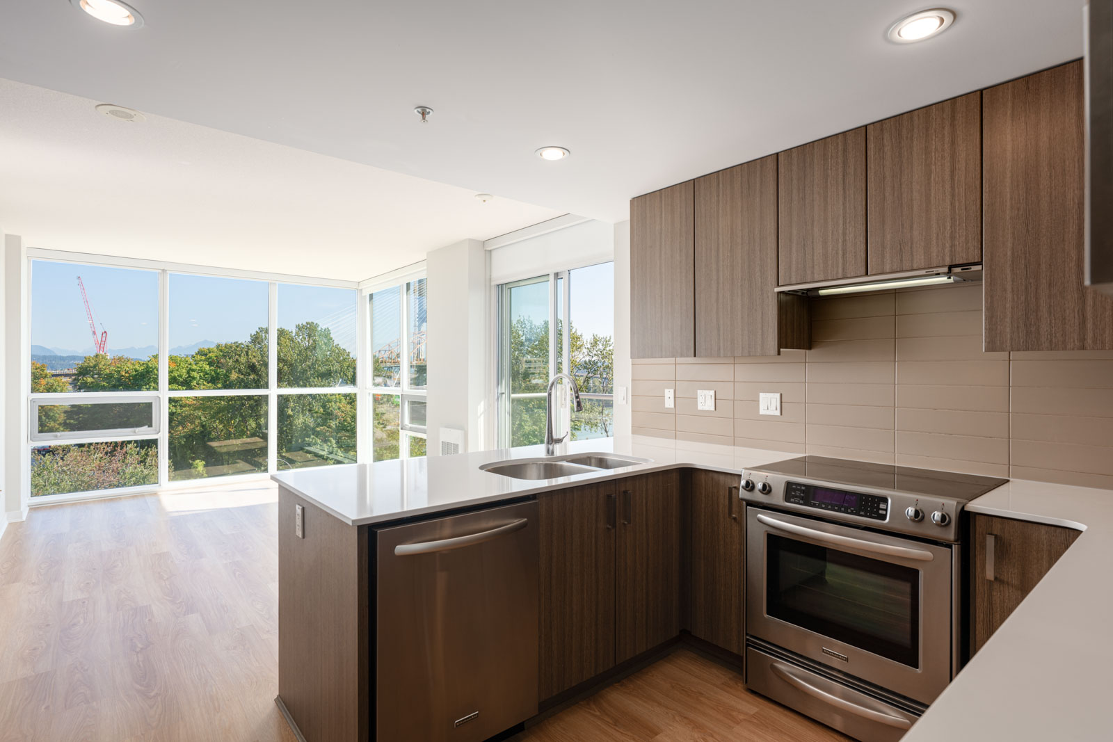 Modern kitchen with wood cabinets, stainless steel appliances, and an island, open to a bright living area with large windows and a view of trees and buildings outside.