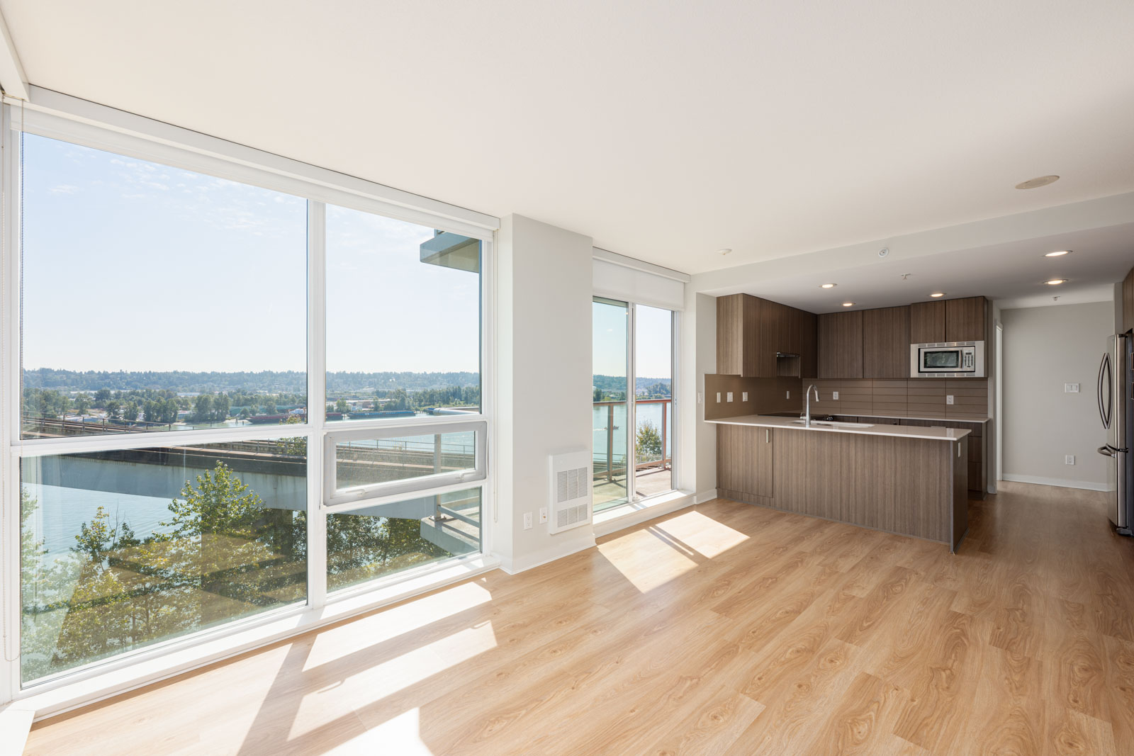 A modern, unfurnished apartment with large windows, light wood floors, an open kitchen, and a view of a river and trees outside.