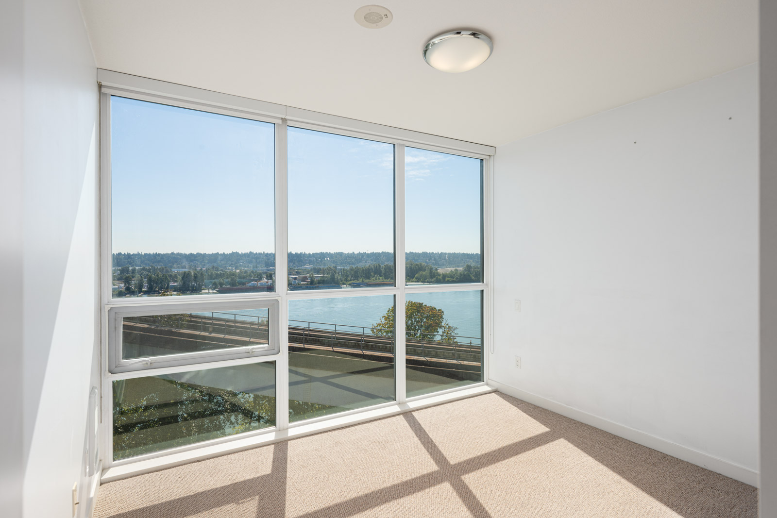 A small empty room with white walls, beige carpet, and large floor-to-ceiling windows offering a view of a river and trees outside.