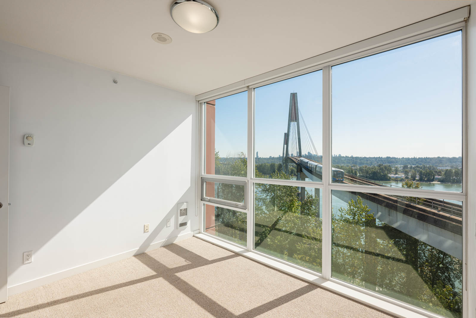 Empty room with large windows showing a view of a cable-stayed bridge, trees, and a river under a clear sky. Sunlight casts shadows on the carpeted floor.