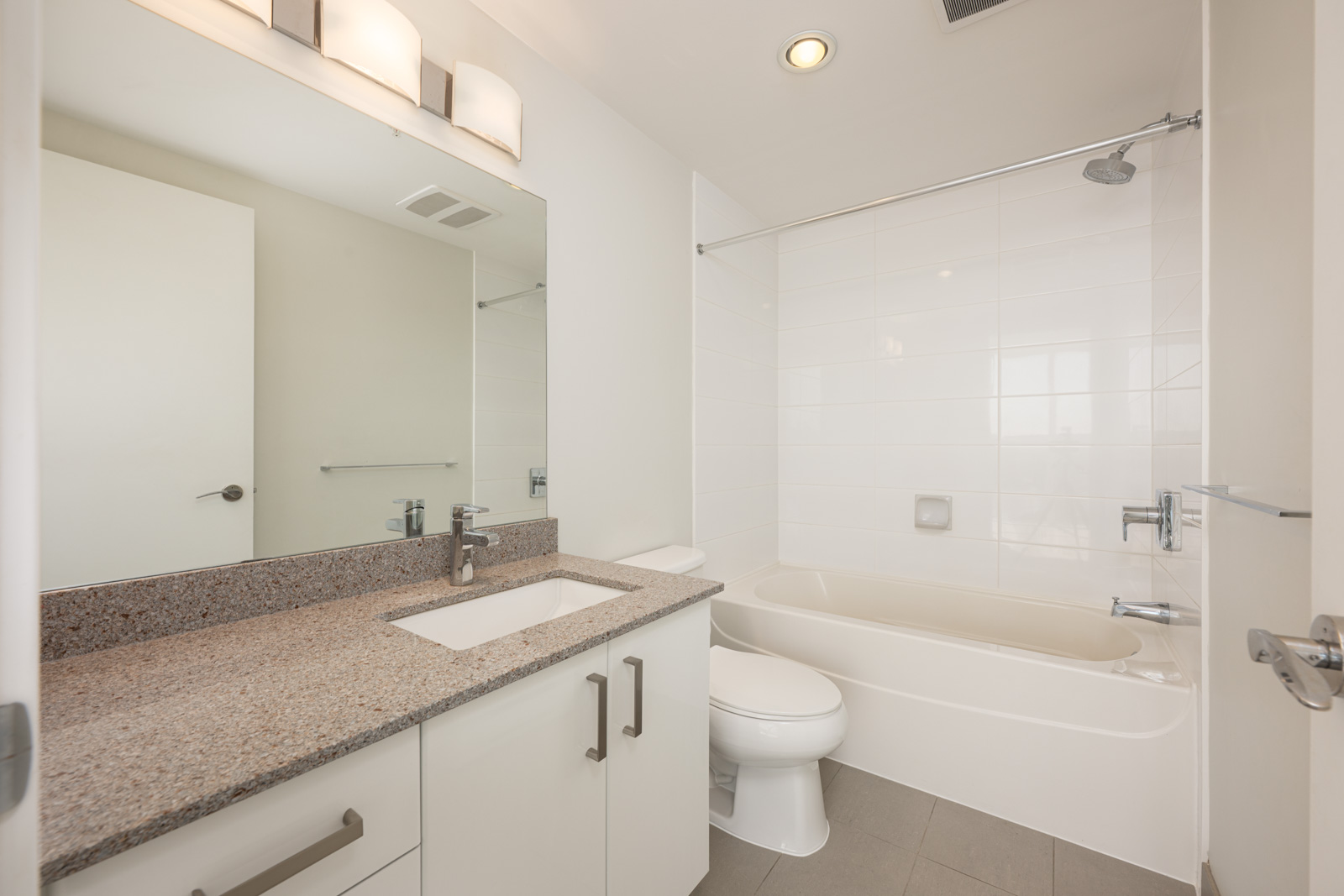 Modern bathroom with a granite countertop, sink, toilet, and a white tiled bathtub-shower combination. Large mirror above the sink and overhead lighting.