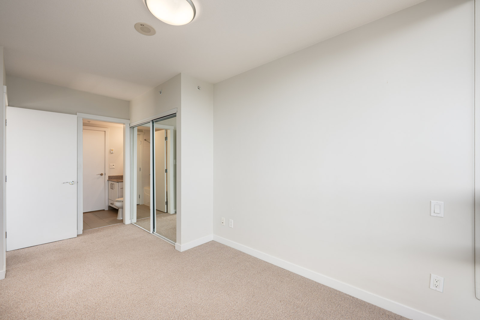 Empty bedroom with beige carpet, white walls, mirrored closet doors, and an open door leading to a bathroom. Ceiling light fixture is visible.