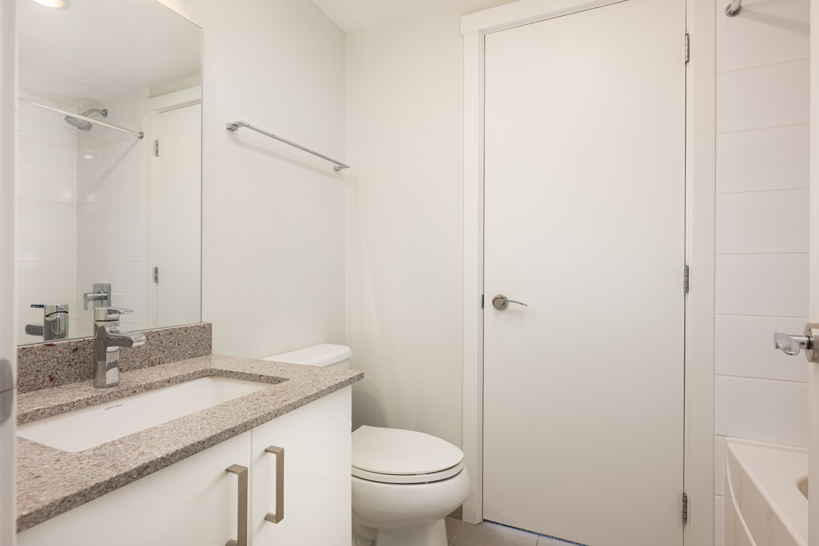 Modern bathroom with white walls, a sink with granite countertop, toilet, towel bar, and a bathtub partially visible on the right.