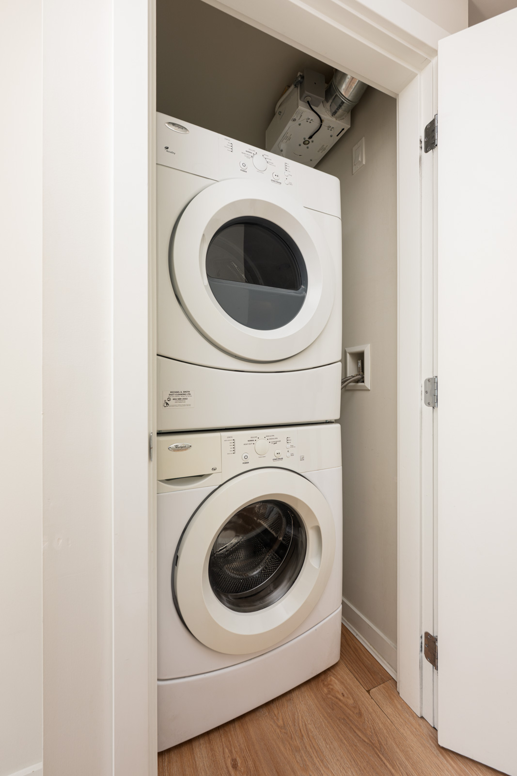 Stacked white front-loading washer and dryer units in a small closet with light wood flooring and white walls.