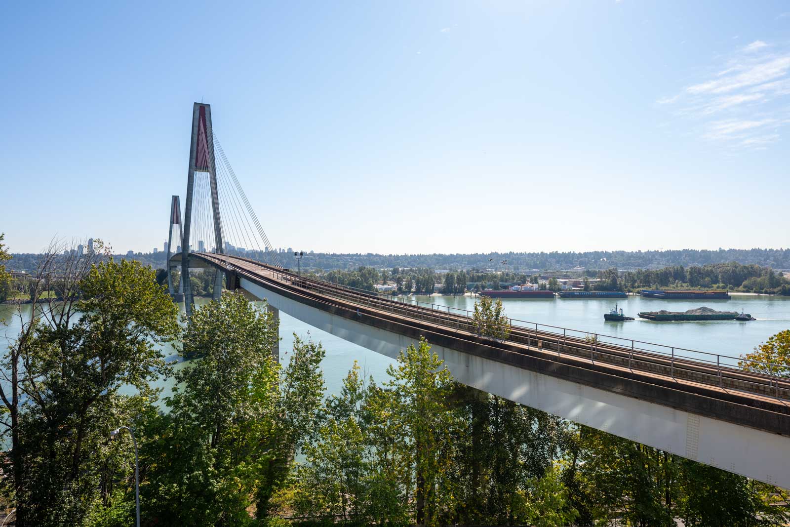 A cable-stayed bridge with a central tower spans a river, with trees in the foreground and a barge on the water below.