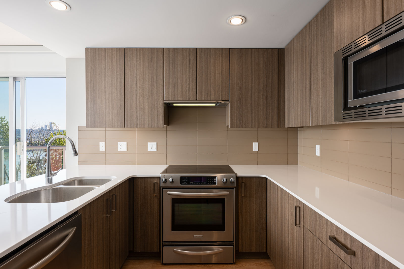 Modern kitchen with wood cabinets, stainless steel appliances, a built-in microwave, double sink, electric stove, and beige tile backsplash under recessed lighting.