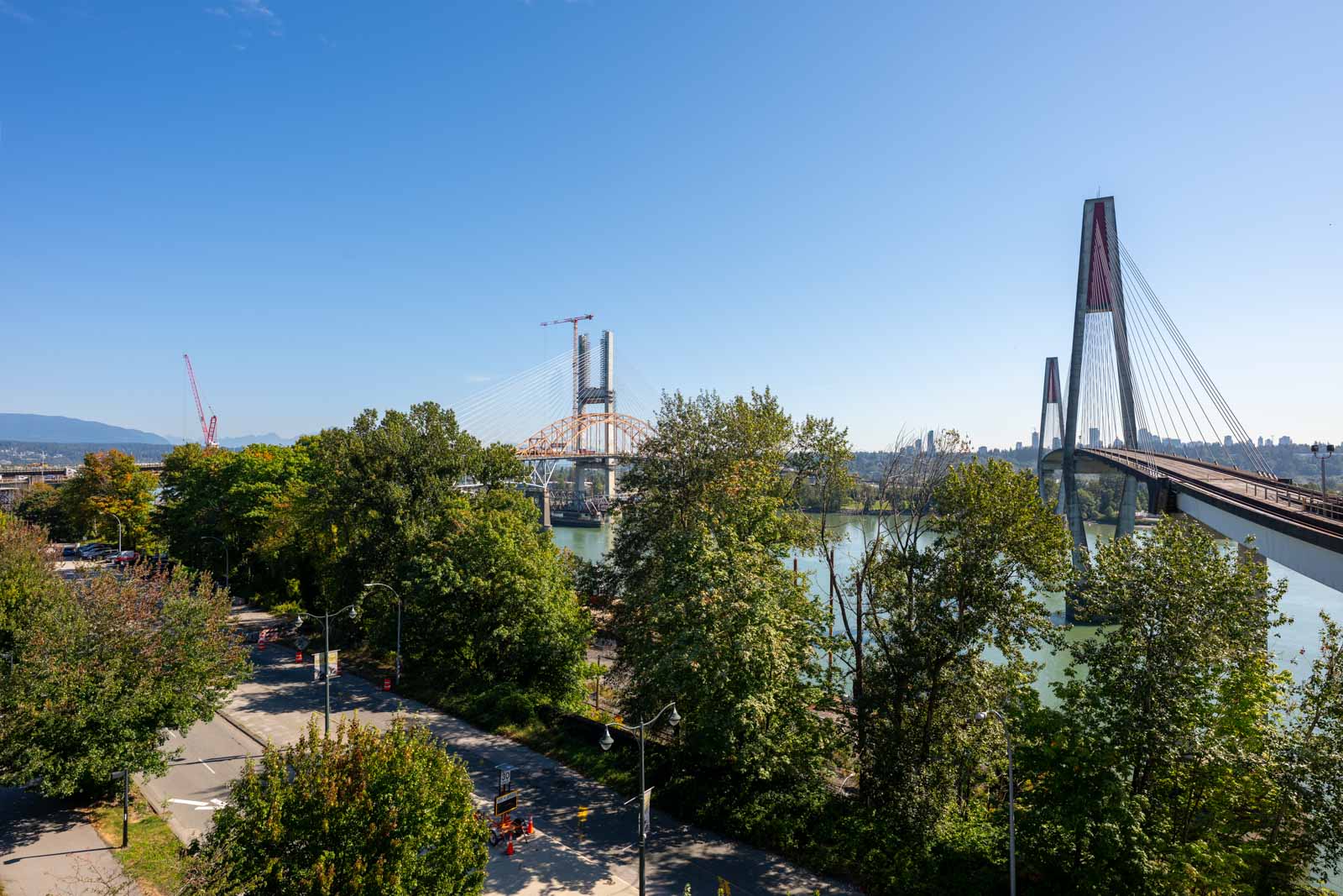 A cable-stayed bridge spans a river, partially obscured by trees, with construction cranes and blue sky in the background.