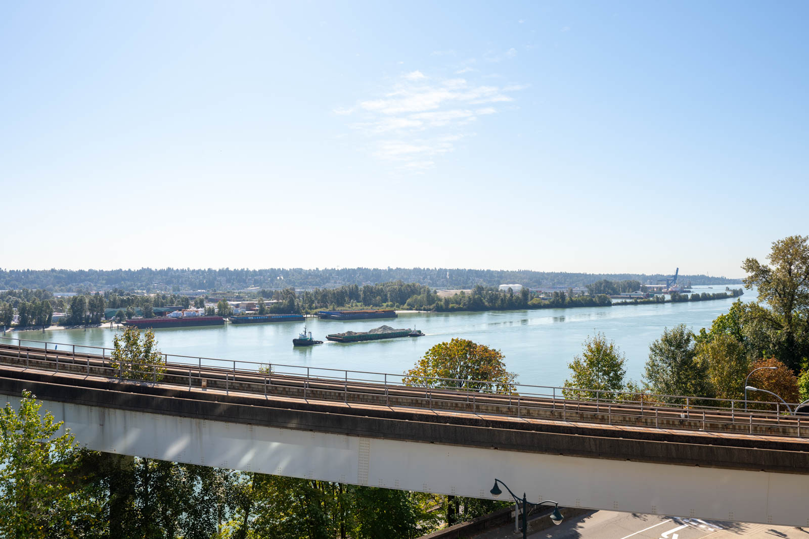 View of a river with cargo barges, trees on both banks, and an elevated roadway in the foreground under a clear, blue sky.