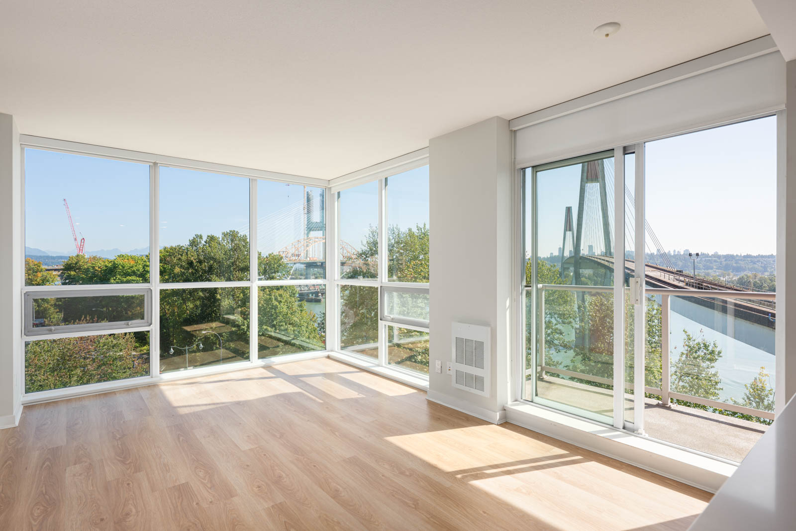 Bright, empty room with large floor-to-ceiling windows, light wood flooring, and views of trees, a river, and a bridge outside.