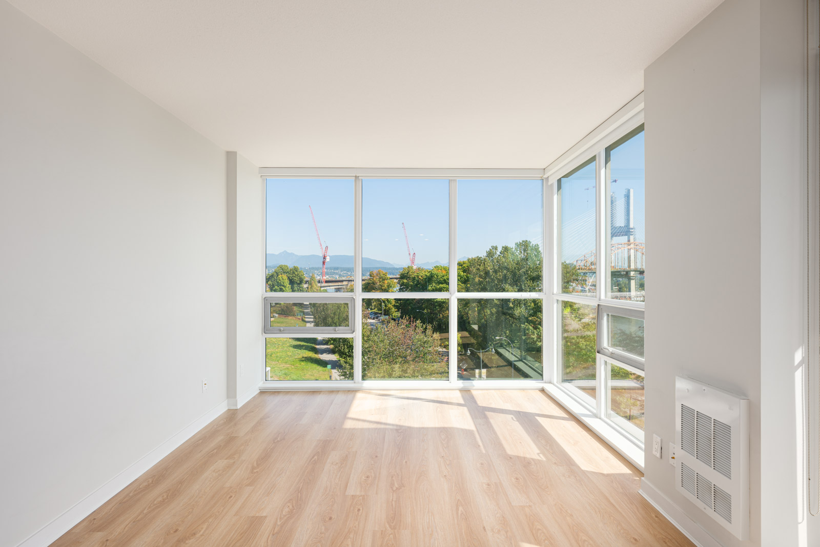 Empty, sunlit room with light wood flooring, white walls, and large floor-to-ceiling windows overlooking a green area, cranes, and a distant bridge.