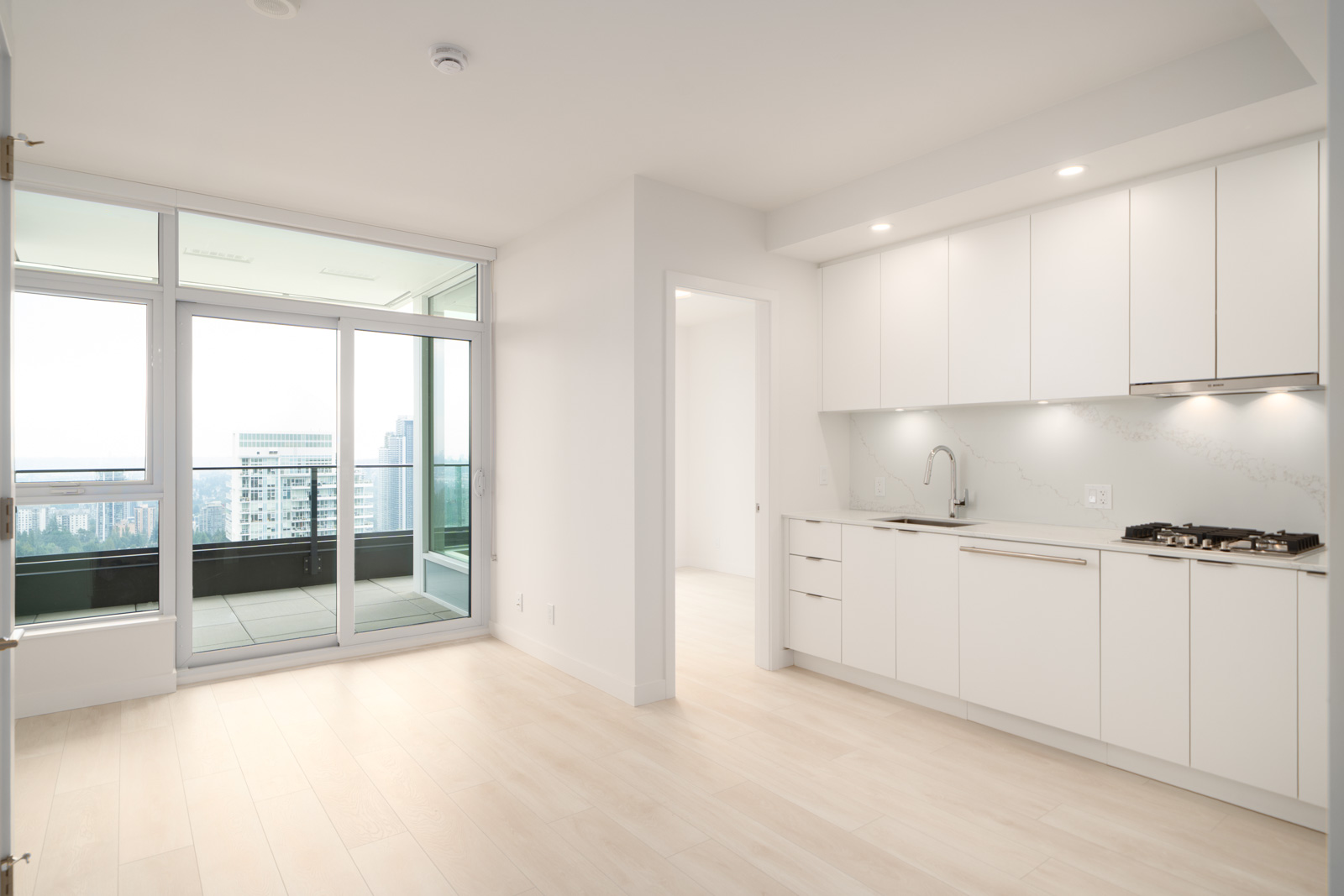 Modern apartment interior with light wood floors, white kitchen cabinets, built-in appliances, and large windows opening to a balcony with a city view.