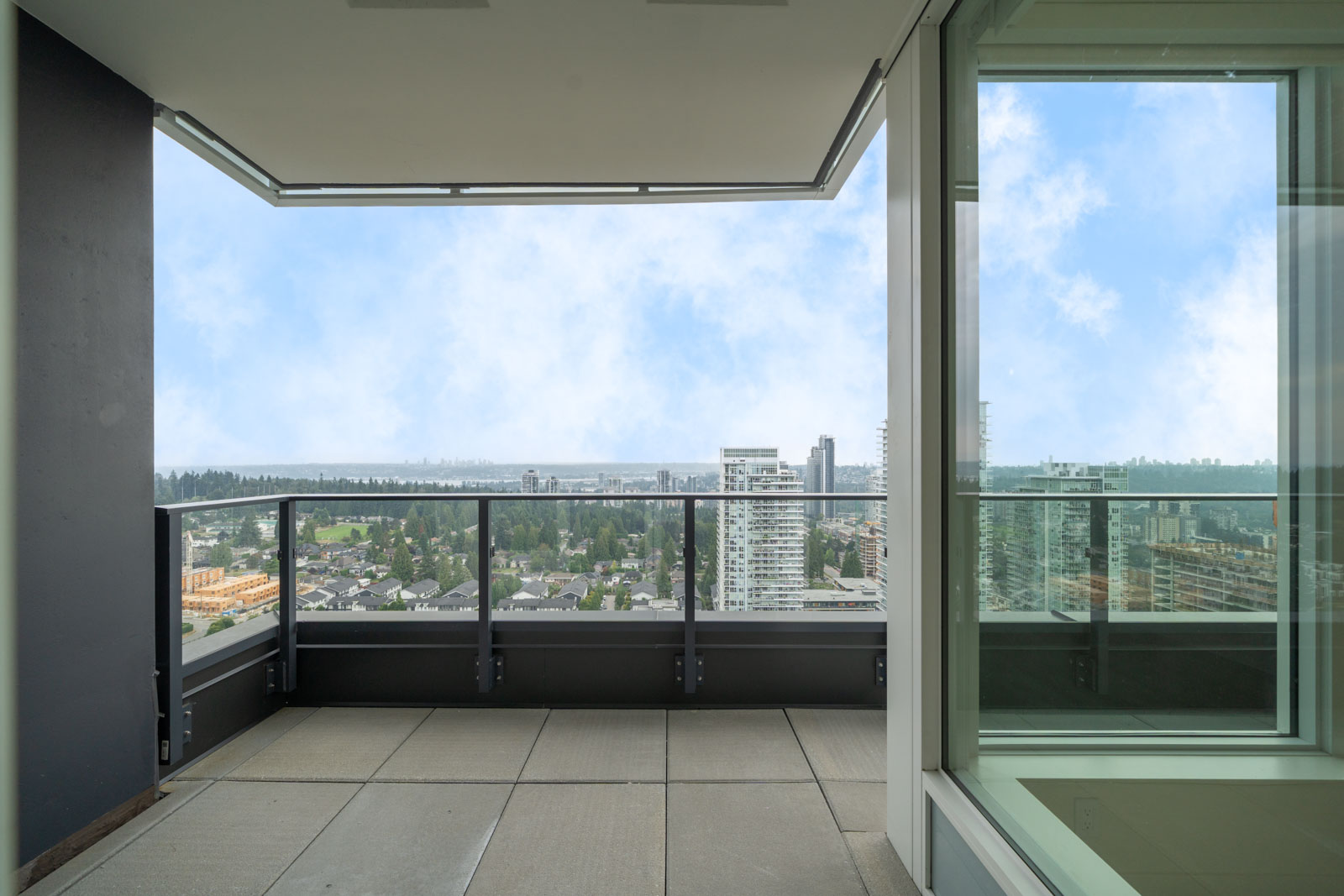 Apartment balcony with glass railing overlooks city buildings and residential area under a partly cloudy sky.