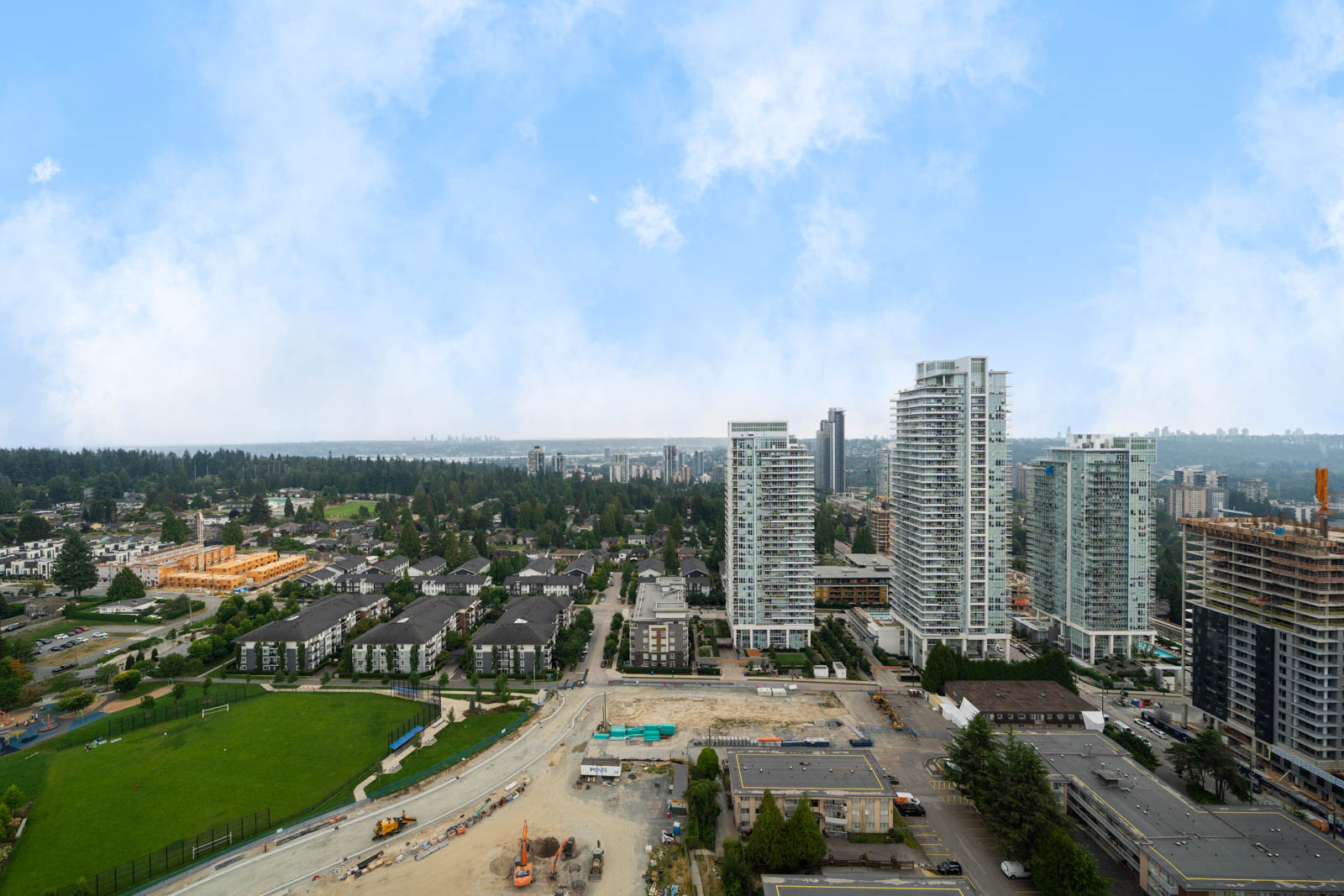 Aerial view of a cityscape with high-rise buildings, mid-rise apartments, a green park, and ongoing construction under a partly cloudy sky.