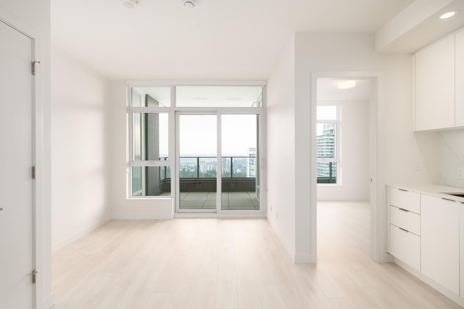Empty modern apartment with light wood floors, large windows, and a glass door leading to a balcony with a city view; partial kitchen and doorway visible on the right.
