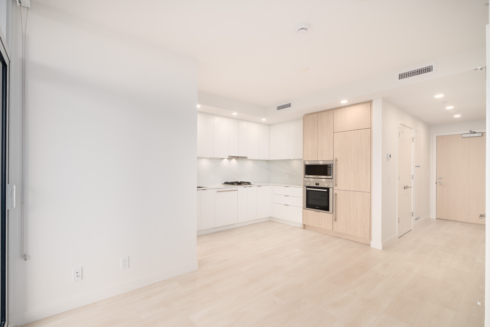 Modern apartment kitchen with white cabinets, built-in stove, oven, and microwave, light wood accents, and light-colored flooring. Entryway door and hallway visible in the background.