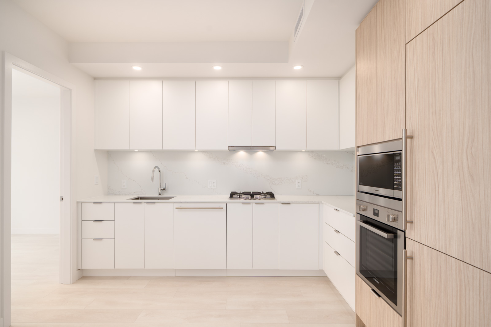 Modern kitchen with white cabinets, light wood accents, built-in oven and microwave, gas cooktop, and under-cabinet lighting.