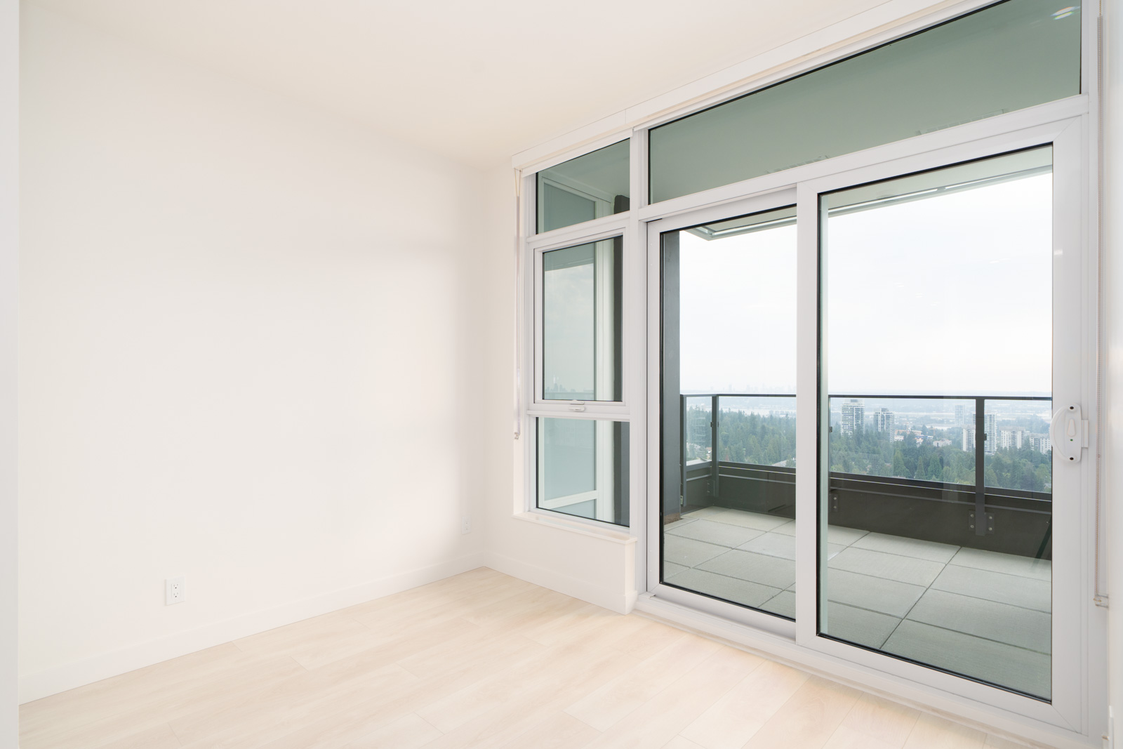 Bright empty room with light wood floors, white walls, and large glass doors leading to a balcony with a city view.