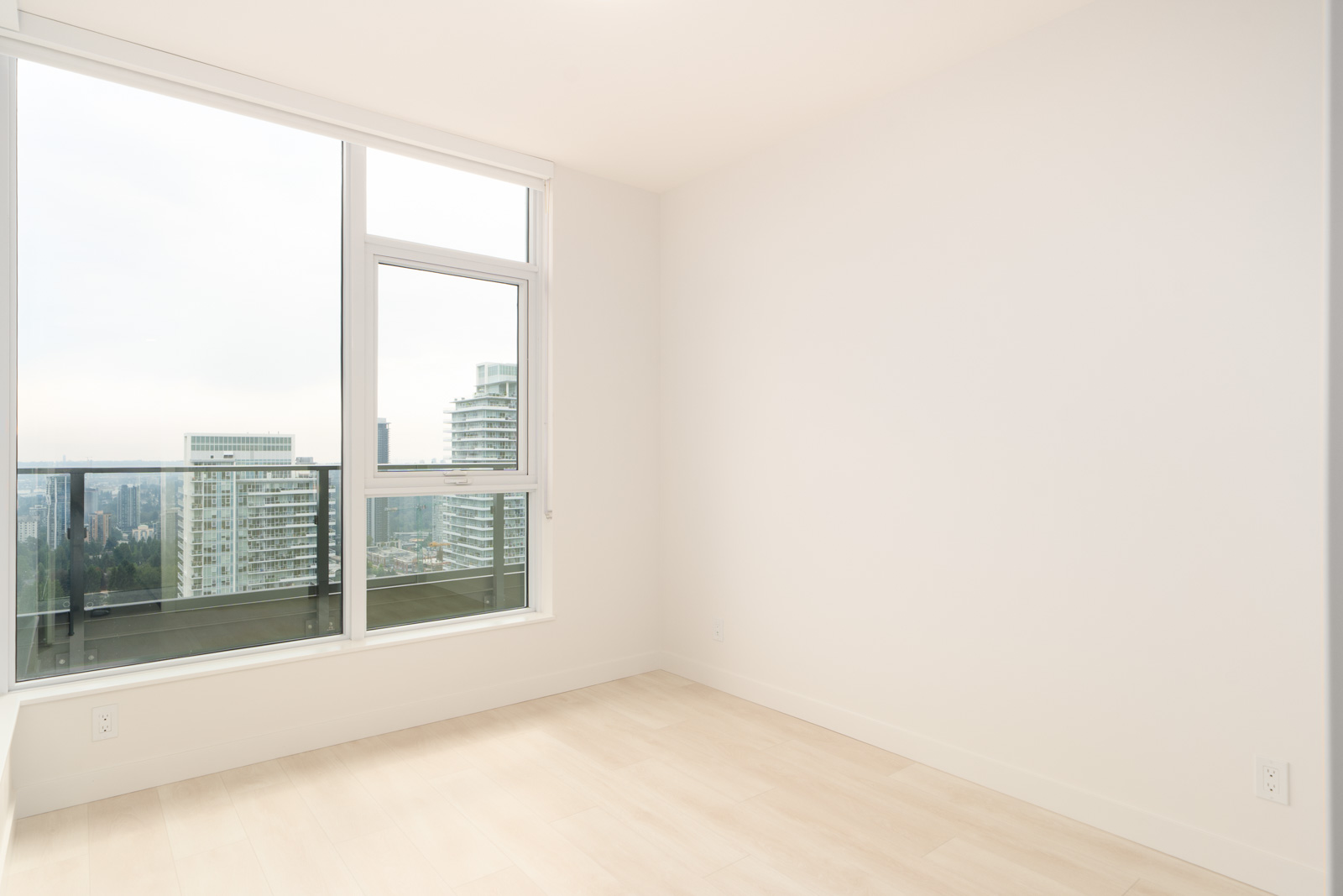 Empty room with light wood flooring, white walls, and a large window overlooking city buildings and a balcony railing.