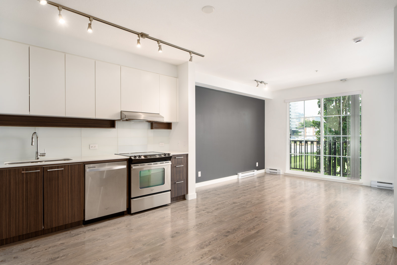 Modern kitchen and living area with wood floors, stainless steel appliances, white cabinets, and large window leading to a balcony with city and tree views.