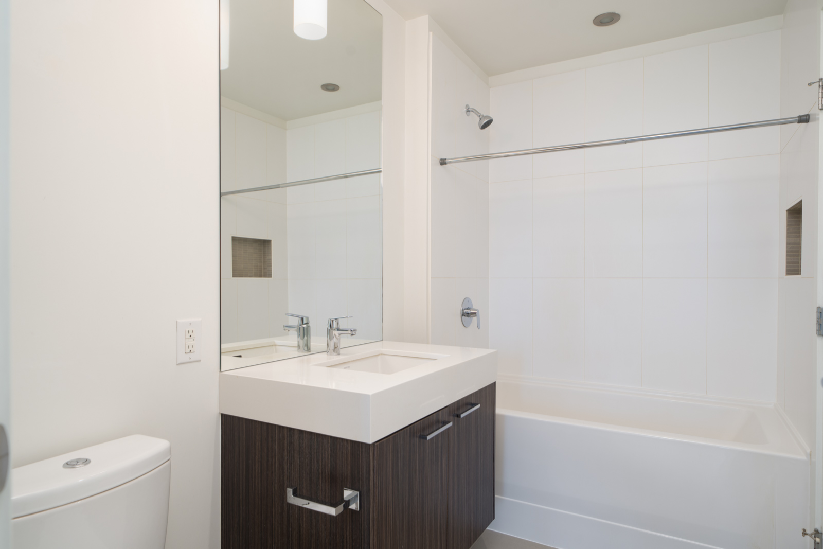 Modern bathroom with white walls, a bathtub with a shower, a large mirror above a sink with a dark wood vanity, and a white toilet in the foreground.