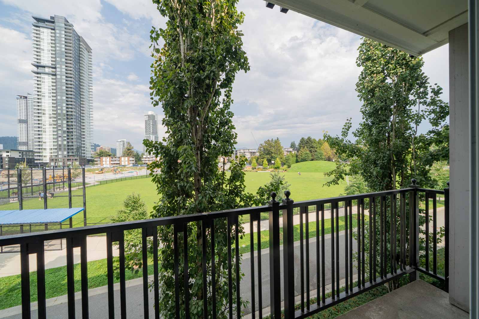 View from a balcony overlooking a park with green grass, trees, and a few tall buildings in the background under a partly cloudy sky.