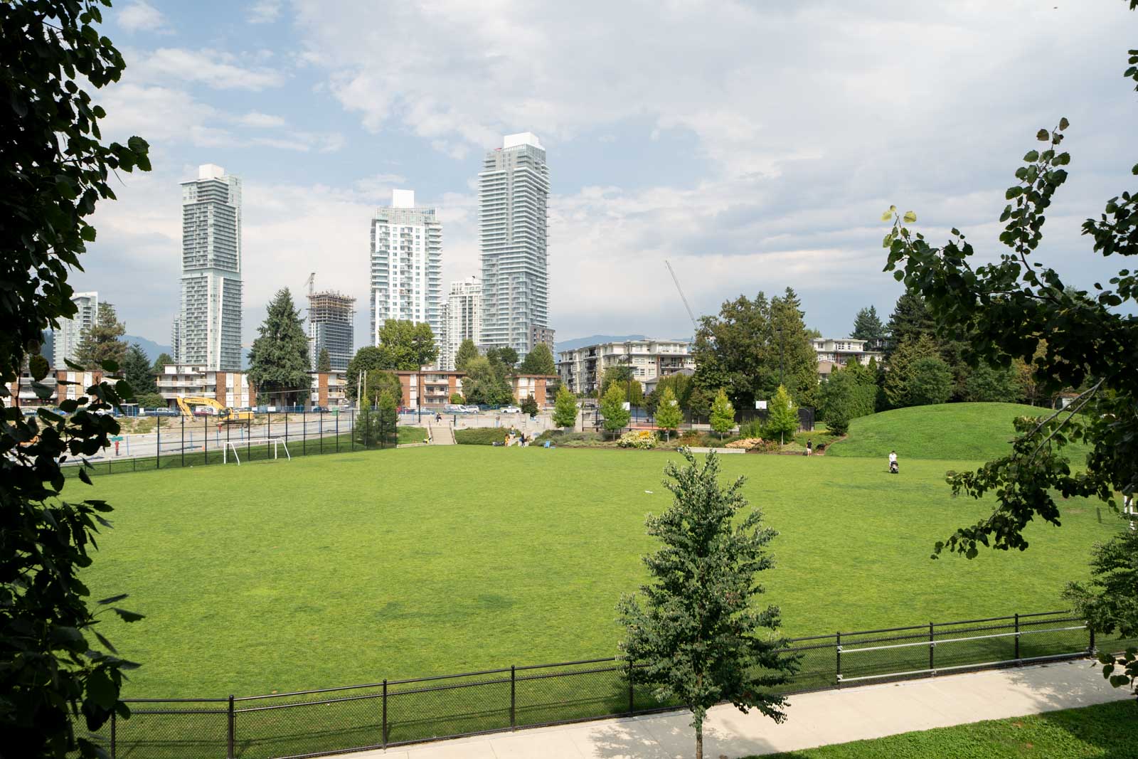 A large grassy park with scattered trees, a sidewalk, and a fence in front; modern high-rise buildings and construction cranes in the background under a partly cloudy sky.