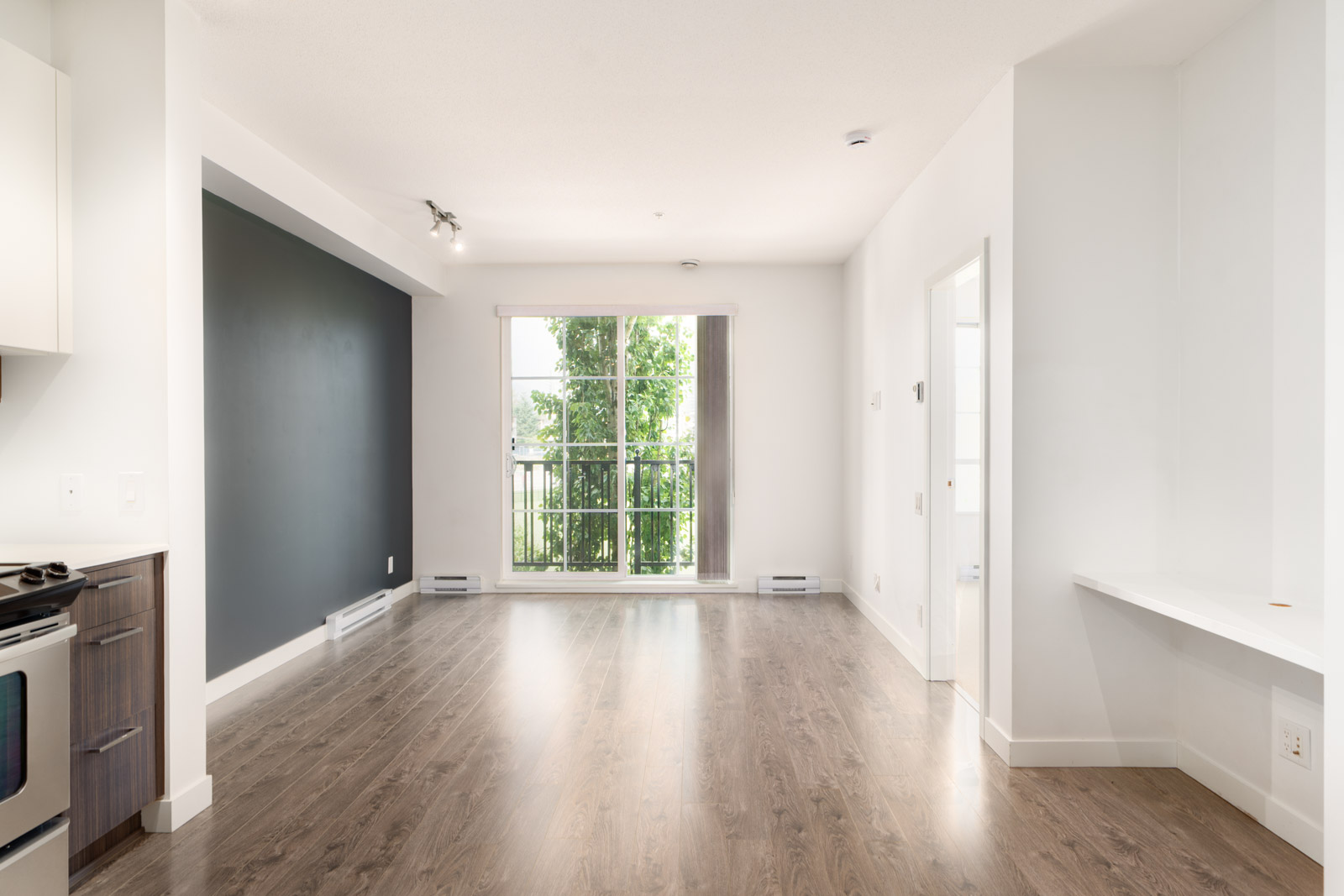 Unfurnished apartment living area with wood flooring, a dark accent wall, large windowed doors opening to a balcony, and a built-in desk on the right.