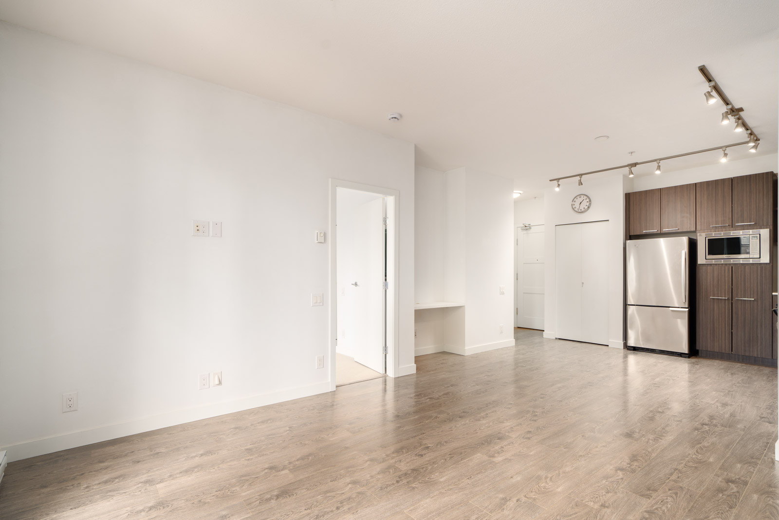 Unfurnished modern apartment with light wood flooring, white walls, stainless steel kitchen appliances, and built-in track lighting.