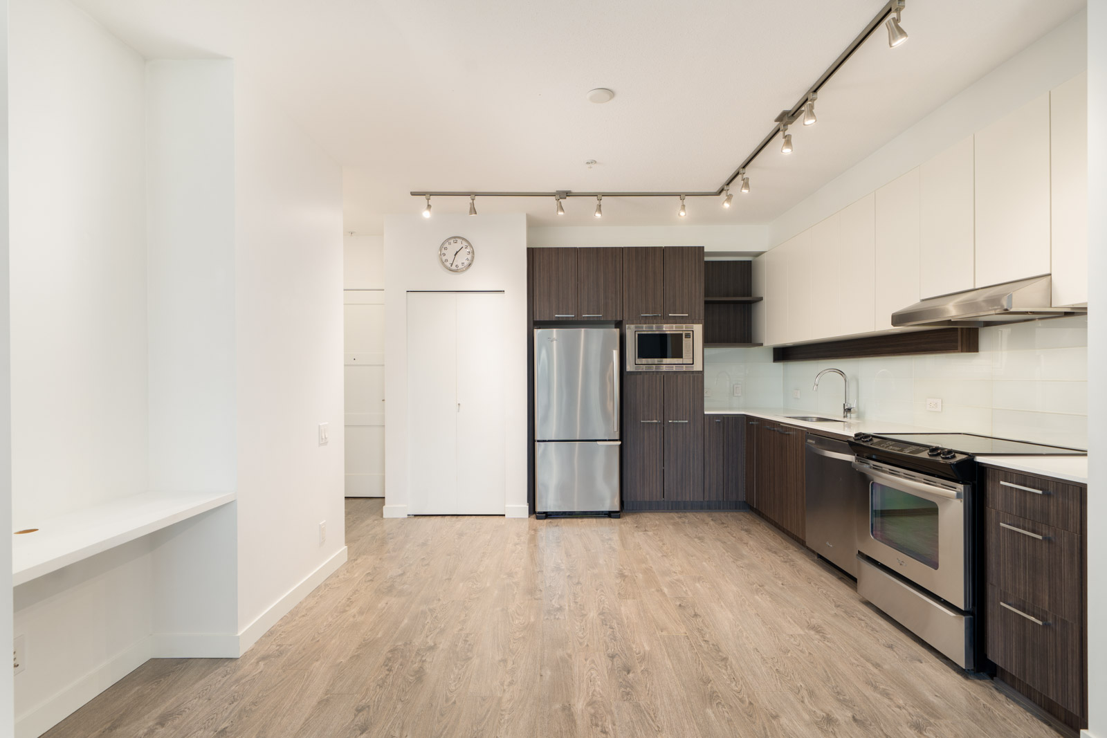 Modern kitchen with stainless steel appliances, wood and white cabinets, track lighting, and light wood flooring. A clock is mounted above a closed door on the left wall.