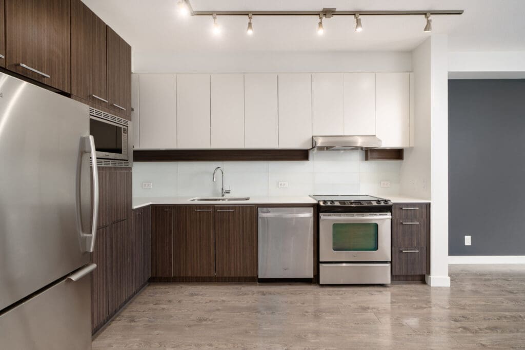 Modern kitchen with stainless steel appliances, white and brown cabinets, a built-in oven, stove, dishwasher, sink, and track lighting above.