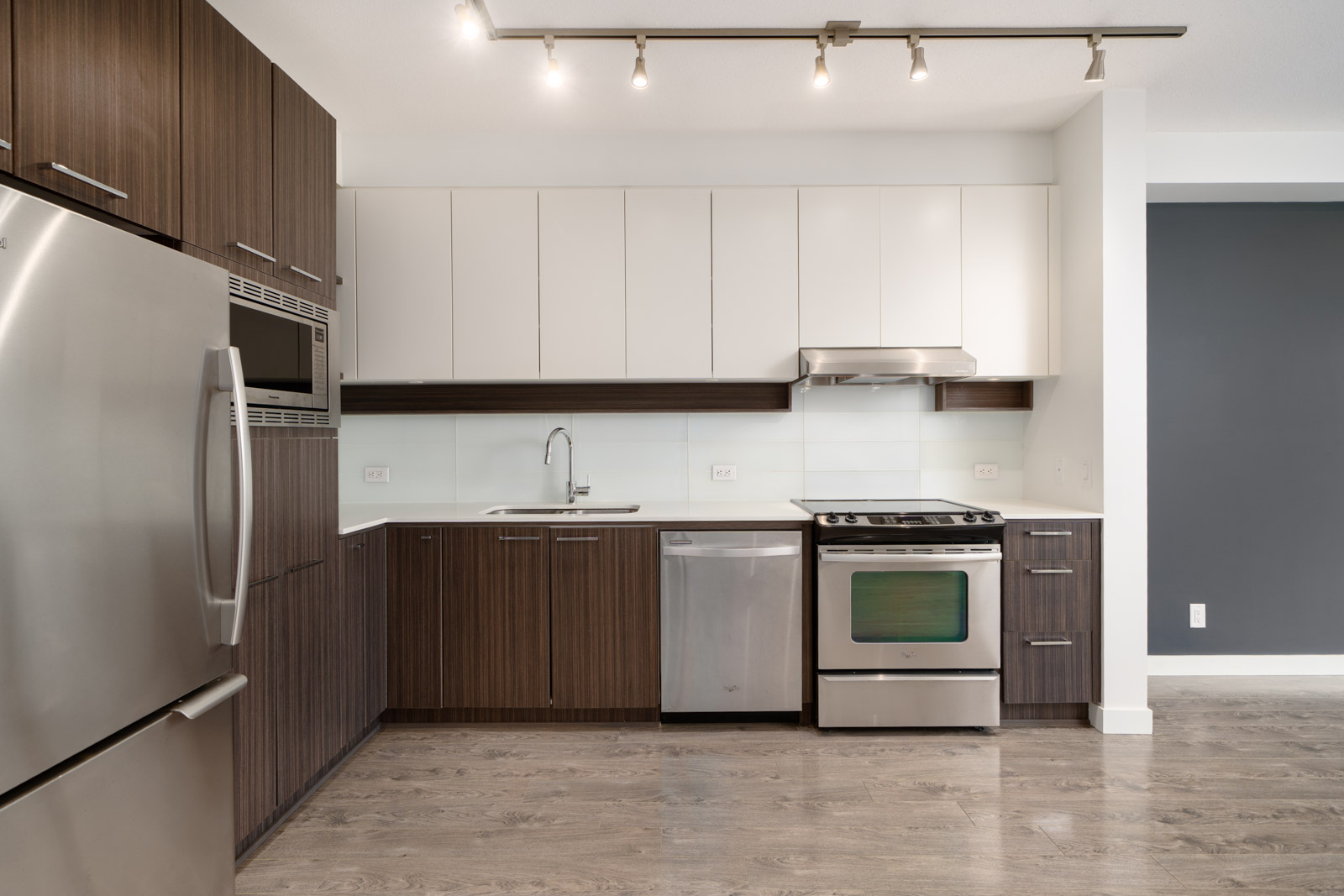 Modern kitchen with stainless steel appliances, white and brown cabinets, a built-in oven, stove, dishwasher, sink, and track lighting above.
