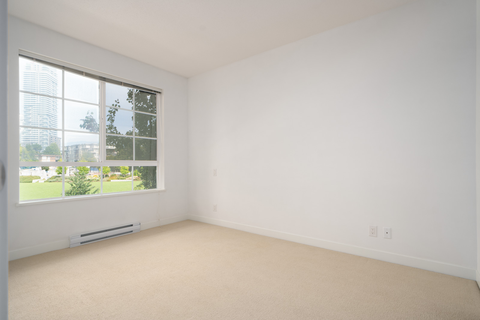 Empty room with beige carpet, white walls, large window showing a cityscape and green lawn, and electrical outlets near the floor.