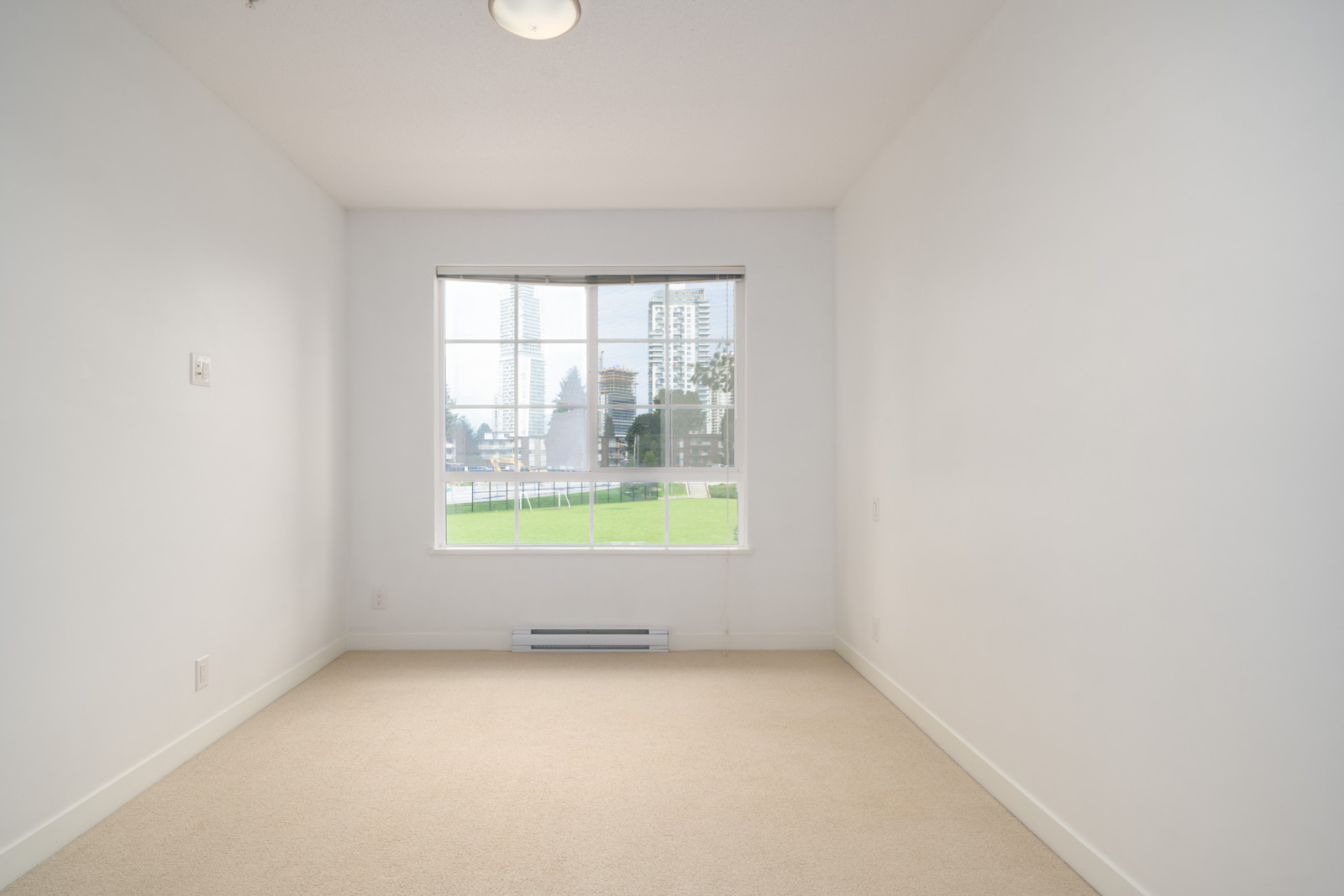 Empty room with white walls, beige carpet, and a large window showing a view of grass and buildings outside.