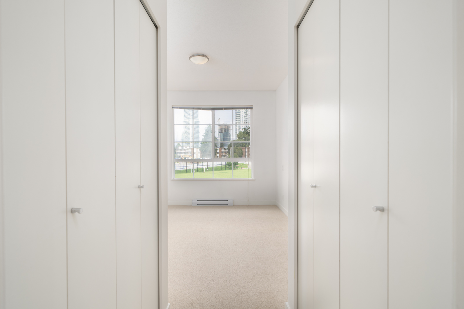 A bright, empty room with beige carpet, white walls, two closets on either side, and a large window showing a partial cityscape and green lawn outside.