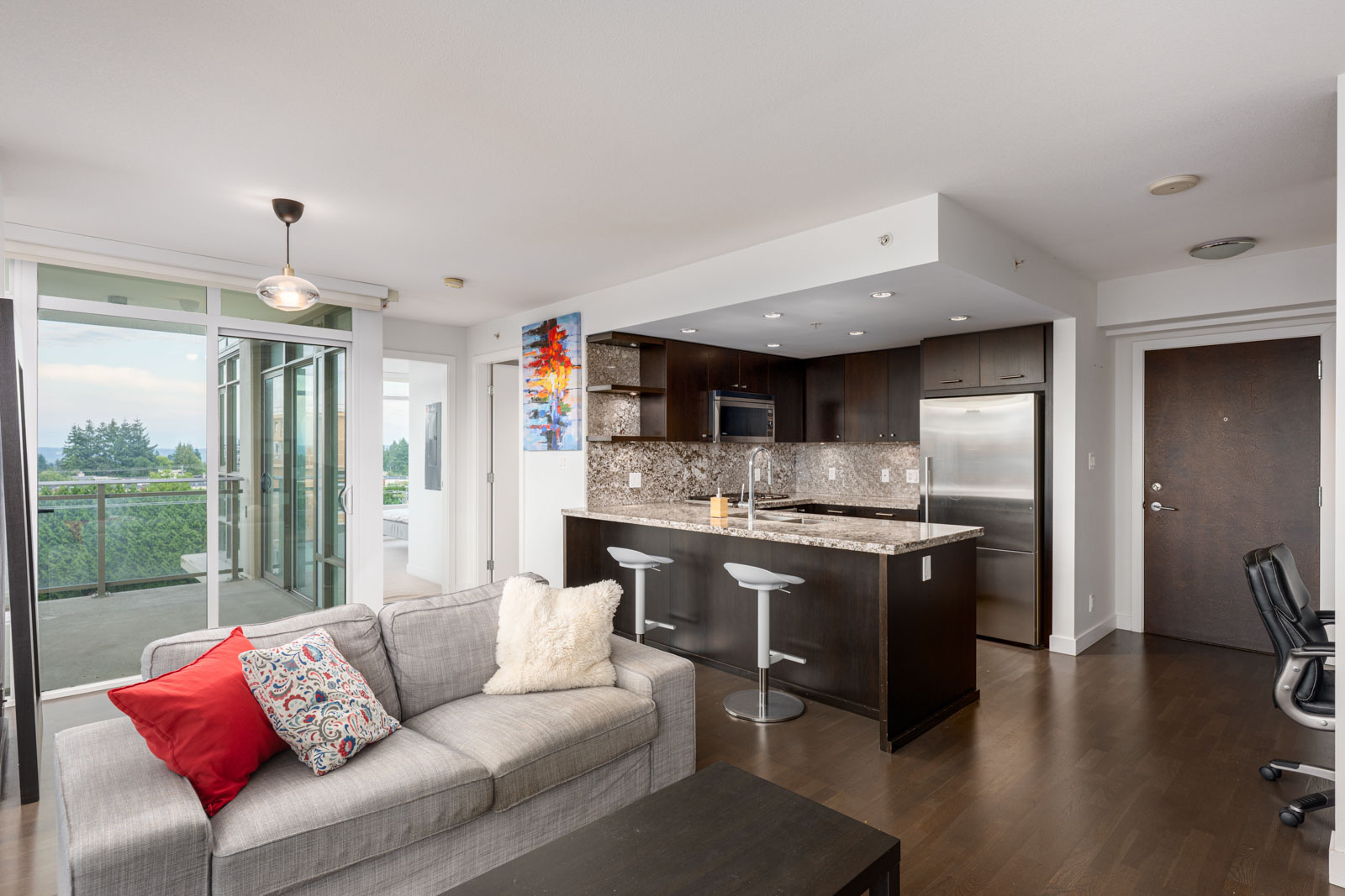 Modern open-plan apartment with a kitchen, island seating, stainless steel appliances, a gray sofa with red and white pillows, and glass doors leading to a balcony with a view.