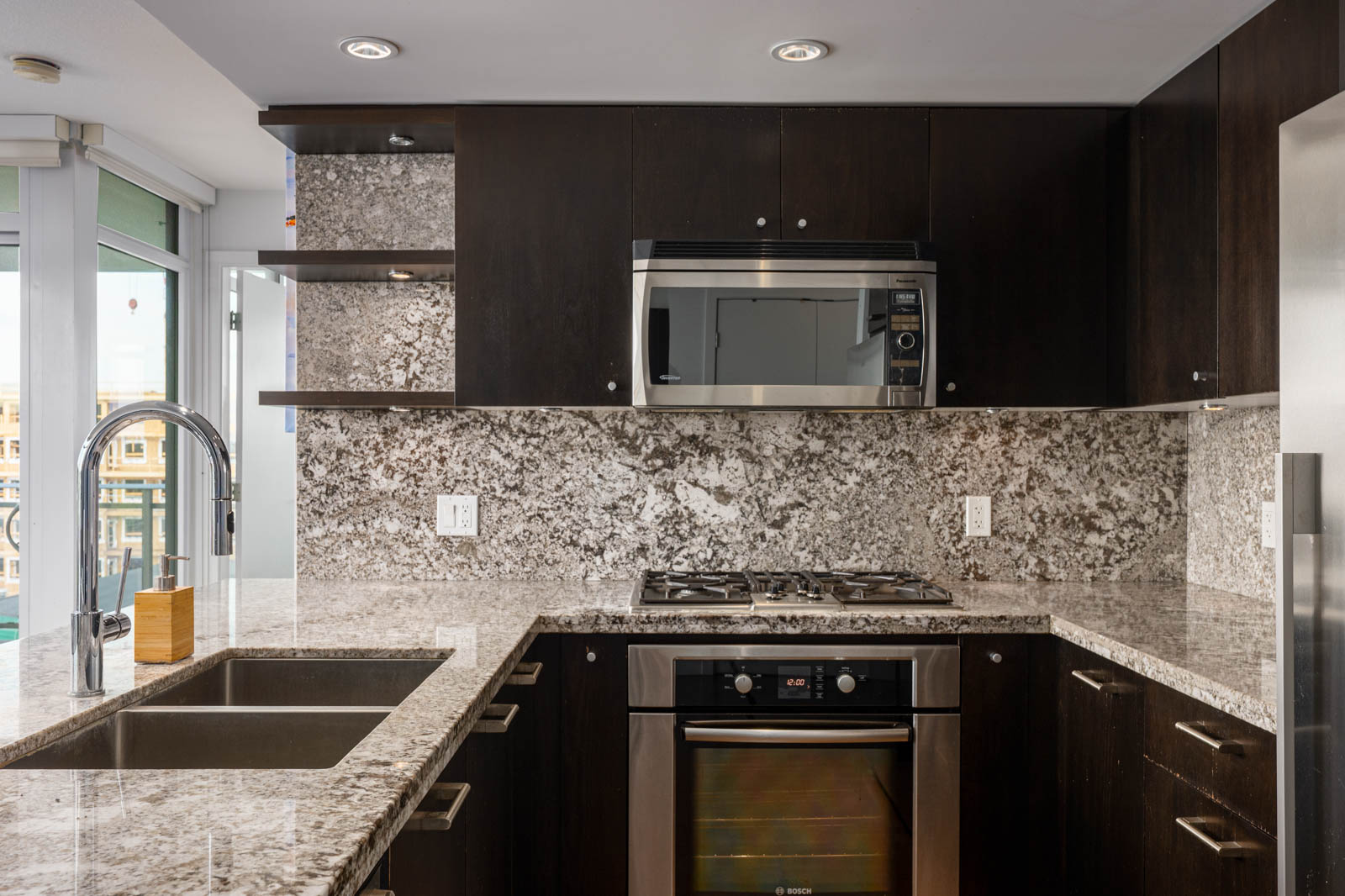 Modern kitchen with dark cabinets, granite countertops and backsplash, stainless steel appliances, and a built-in gas stove beneath a microwave.