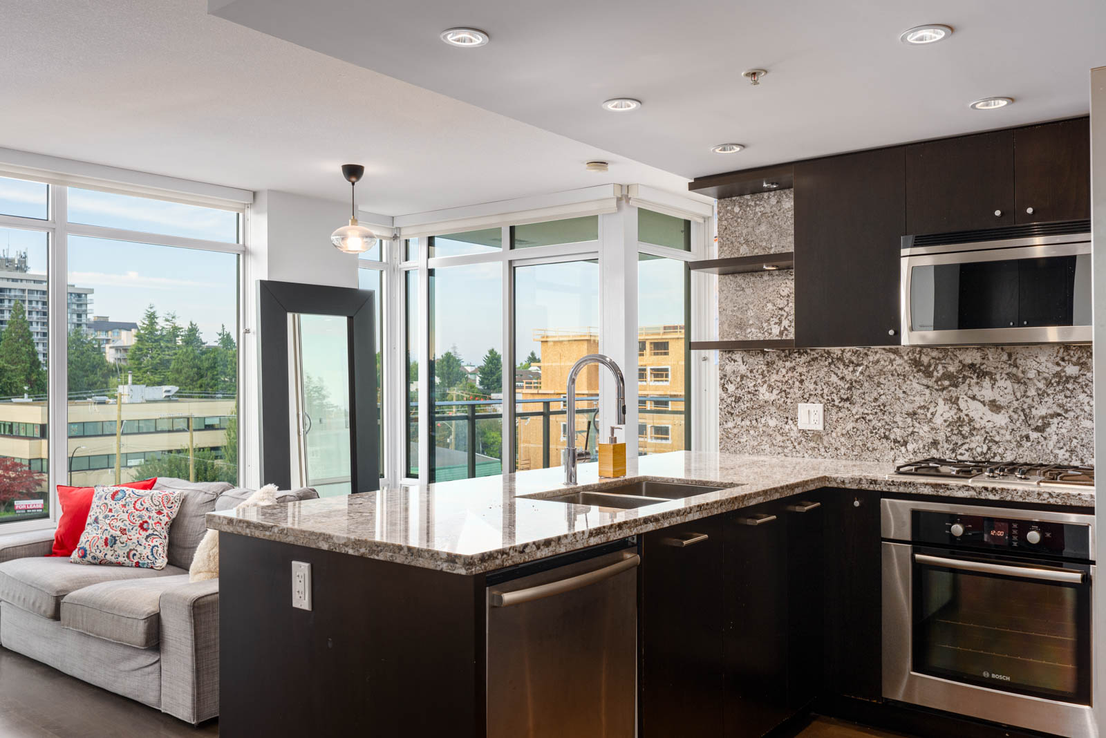 Modern kitchen with dark cabinets, granite countertops, built-in stainless steel appliances, and a large window overlooking a cityscape. A gray sofa with colorful pillows is nearby.