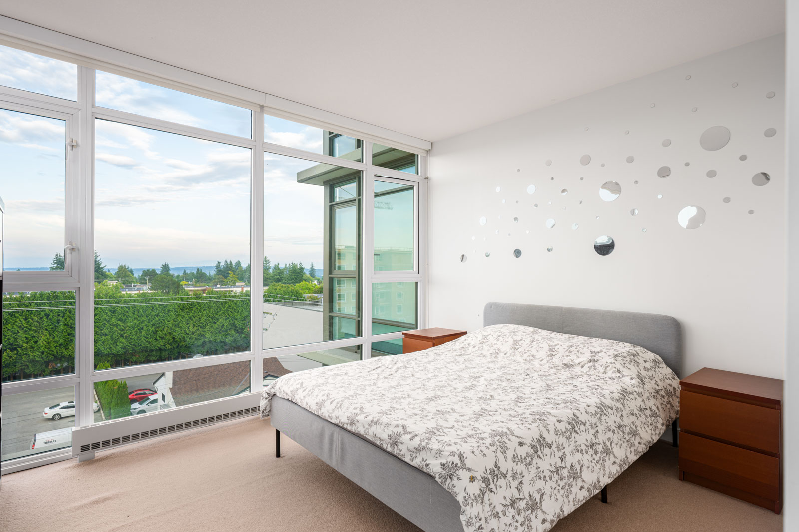 A bright bedroom with large floor-to-ceiling windows, a gray bed with a white patterned duvet, two wooden nightstands, and circular wall decals above the bed.