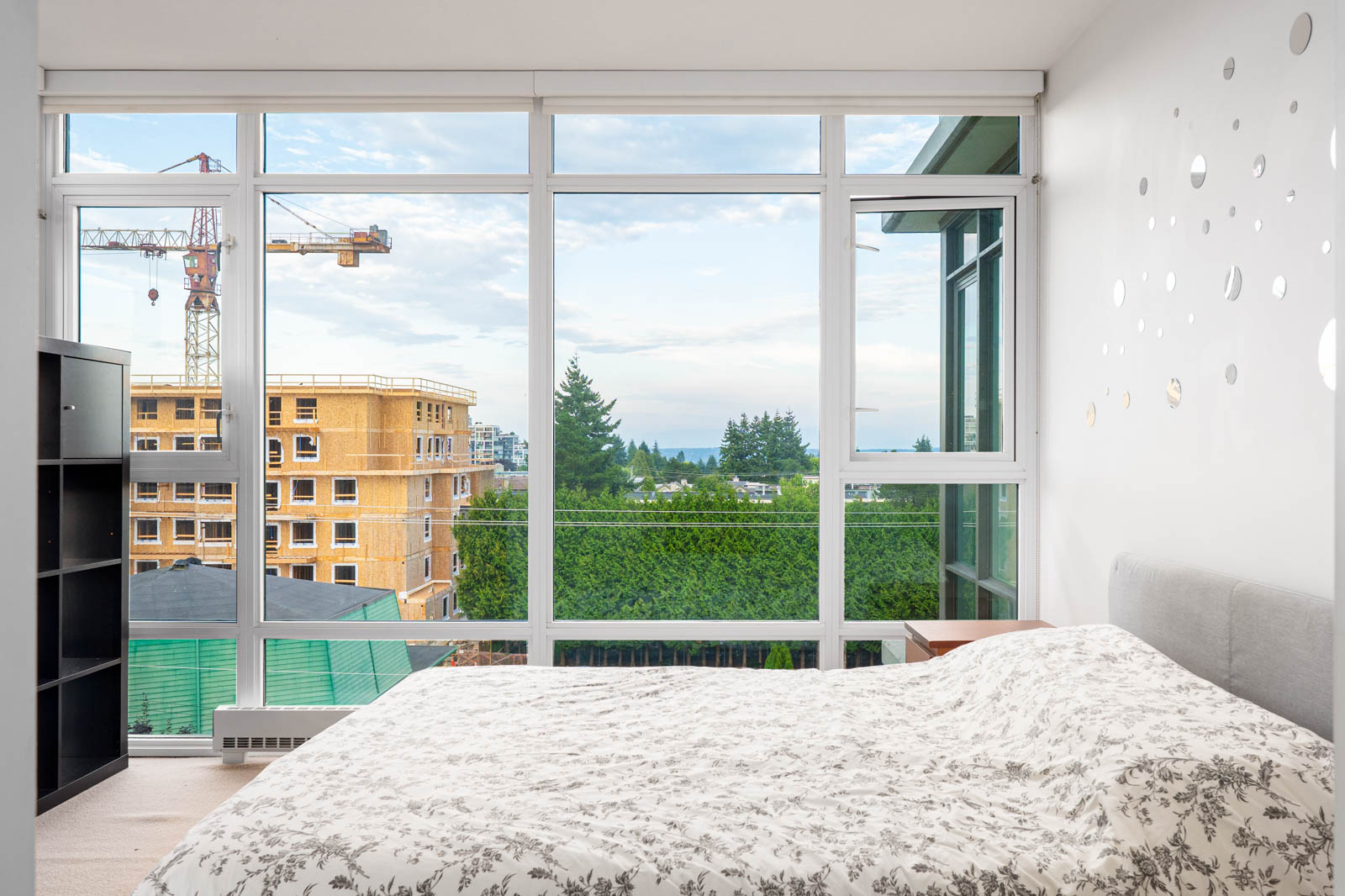 Bedroom with large windows, a bed with a patterned cover, and a view of construction, trees, and the sky outside.