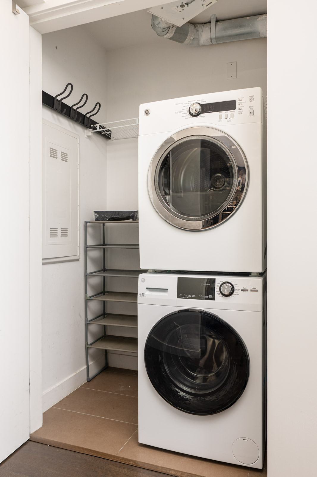 A stacked washer and dryer in a small laundry closet with a shelving unit and wall hooks on the left side.
